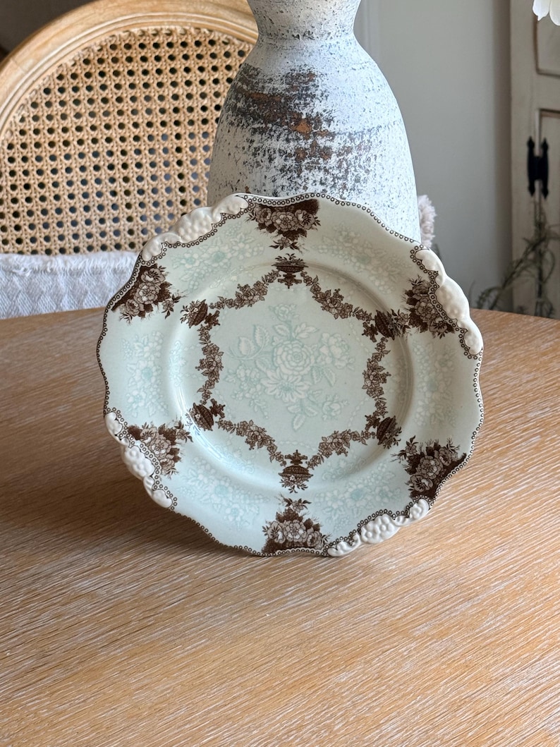 May include: A vintage porcelain plate with a scalloped edge. The plate features a light blue background with a brown floral pattern around the rim and center. The plate is displayed on a light wood surface.