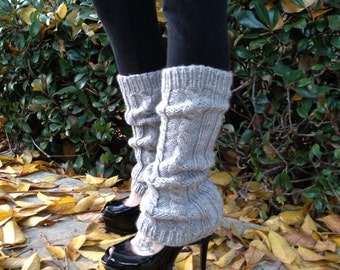 May include: Gray knitted leg warmers with a cable knit pattern worn with black high-heeled shoes. The shoes have a peep toe and a platform sole. The person is standing on a bed of fallen autumn leaves.