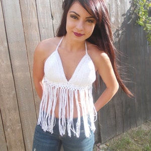 May include: White crochet halter top with fringe detailing. The top is worn with blue jeans.