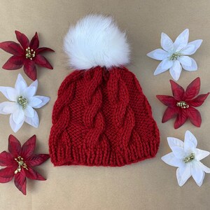 May include: Red knitted beanie with a white pom pom. The beanie has a cable knit pattern and is surrounded by red and white poinsettia flowers.