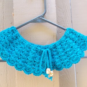 May include: A teal blue crocheted collar with a scalloped edge and a tie closure. The collar is decorated with small white beads.