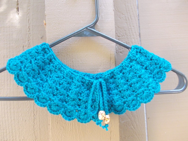 May include: A teal blue crocheted collar with a scalloped edge and a tie closure. The collar is decorated with small white beads.
