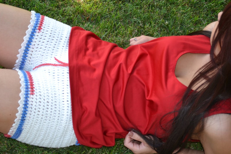 May include: A white crocheted shorts with red and blue trim, worn with a red sleeveless top. The person is lying on green grass.