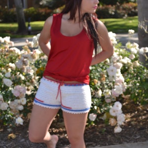 May include: A woman wearing a red tank top and white crochet shorts with blue and red trim. She is standing in front of a bed of white flowers.