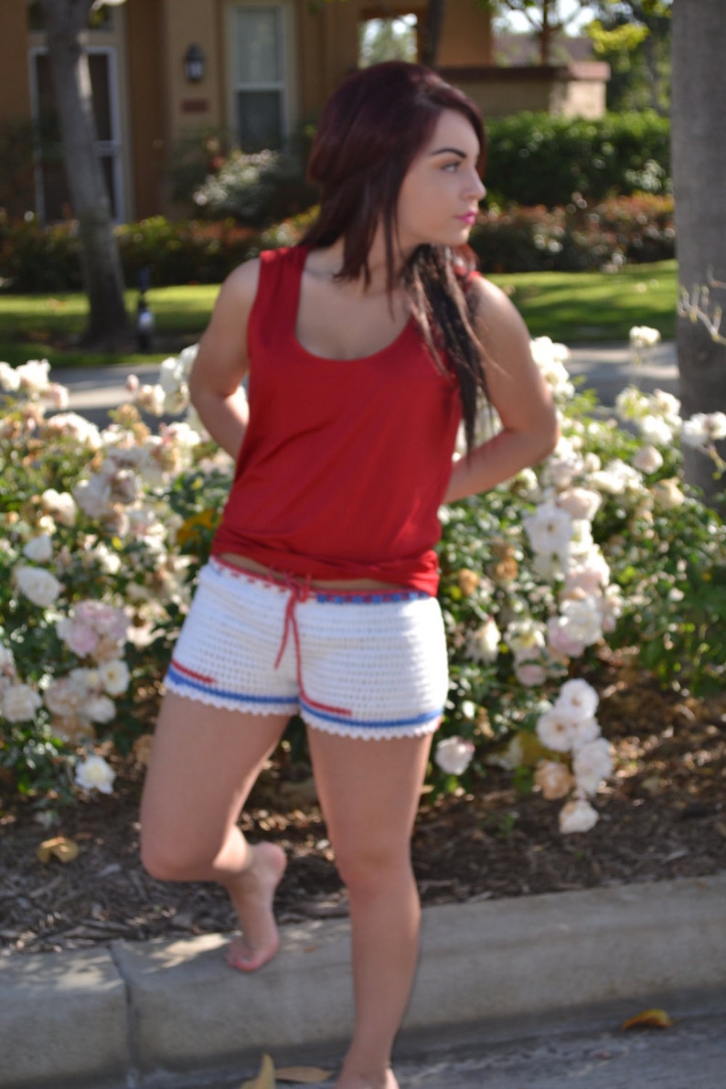May include: A woman wearing a red tank top and white crochet shorts with blue and red trim. She is standing in front of a bed of white flowers.