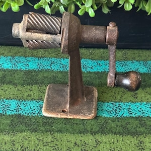 May include: An antique, cast-iron pencil sharpener with a hand crank. The sharpener is a dark, rusty brown color and has a rectangular base. The crank has a wooden handle. The blades are visible inside the sharpener.