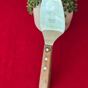 May include: A stainless steel pie server with a wooden handle. The server has a wide, flat blade with the text "kai cut" and other markings. The handle is attached with three rivets.