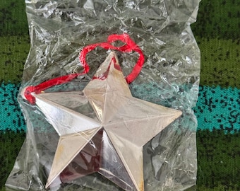 May include: A silver star-shaped decoration with a red hanging ribbon, packaged in a clear plastic bag. The star has five points and a metallic finish, suitable for festive occasions.