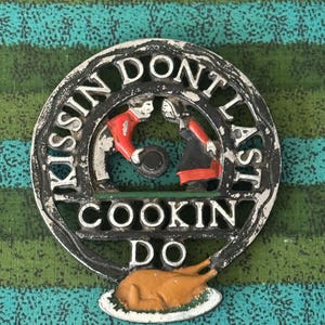 May include: A circular cast iron trivet featuring a distressed black finish with silver lettering that reads KISSIN DONT LAST COOKIN DO. The design includes two figures in red and black clothing and a roasted brown turkey at the bottom base.