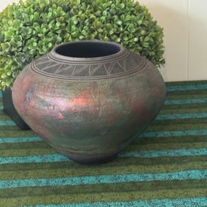 May include: A round ceramic vase with a wide opening and a dark, textured finish. The surface features a mottled blend of deep green, copper, and black tones, with a decorative band of etched triangular patterns encircling the neck area.