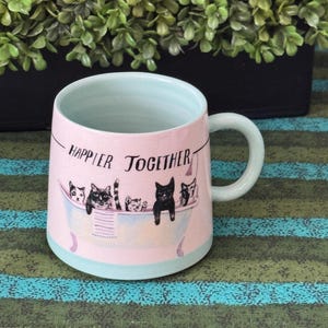 May include: Pink ceramic mug with a light blue interior and handle. The mug features a design of cats in a bathtub with the words "Happier Together" printed on the front. The mug is on a striped surface.