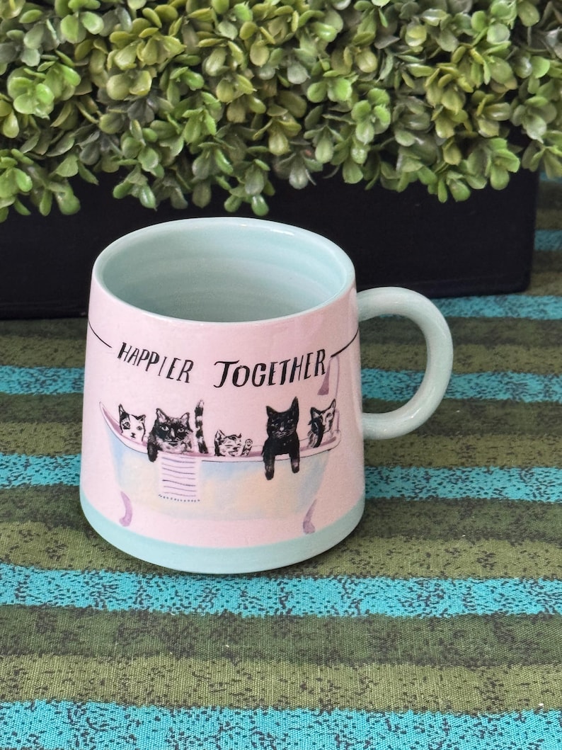May include: Pink ceramic mug with a light blue interior and handle. The mug features a design of cats in a bathtub with the words "Happier Together" printed on the front. The mug is on a striped surface.