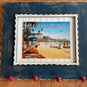 May include: A blue wooden frame with a white scalloped edge surrounds a vintage-style print of a beach scene. The print features the text "Waikiki Beach" and depicts people relaxing on the sand. The frame has four red knobs on the bottom.