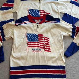 May include: Two white hockey jerseys with red, white and blue stripes. The jerseys feature a graphic of an American flag with the word "USA" printed below it.