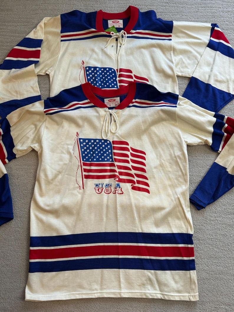 May include: Two white hockey jerseys with red, white and blue stripes. The jerseys feature a graphic of an American flag with the word "USA" printed below it.