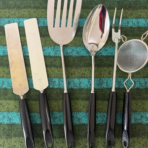 May include: A set of six stainless steel kitchen utensils with black handles. The set includes a spatula, two offset spatulas, a slotted turner, a spoon, a fork, and a strainer.
