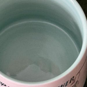 May include: A ceramic bowl with a light blue interior and a pink exterior. The exterior features the words "Happier Together" in black script, along with illustrations of dogs and cars. The bowl has a smooth, glossy finish.