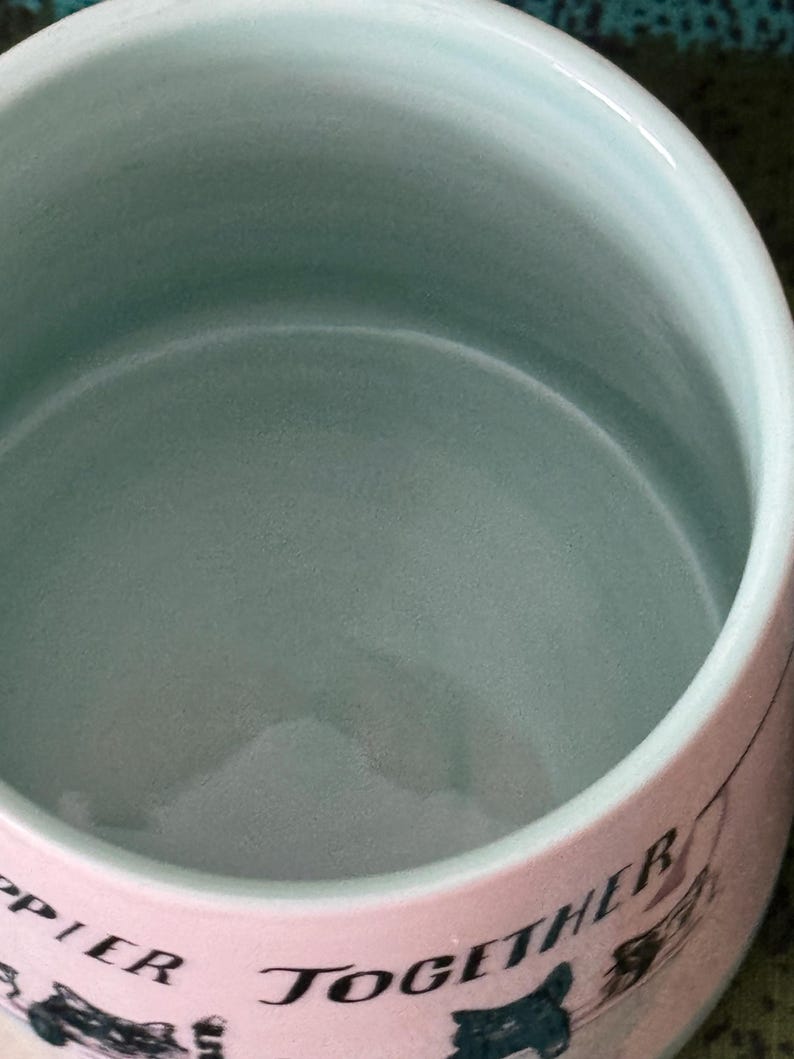 May include: A ceramic bowl with a light blue interior and a pink exterior. The exterior features the words "Happier Together" in black script, along with illustrations of dogs and cars. The bowl has a smooth, glossy finish.