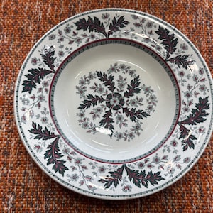 May include: White ceramic bowl with a floral design. The bowl features a dark green and burgundy leaf pattern around the rim and a central floral motif. The design is set against a white background.