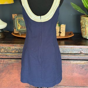 Pu&ograve; includere: Abito senza maniche blu navy con tessuto strutturato. Lo scollo e le spalline sono rifiniti con una fascia decorativa dorata e color crema. L'abito &egrave; esposto su un manichino nero.