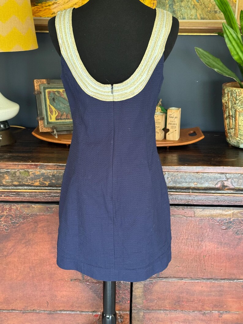 Pu&ograve; includere: Abito senza maniche blu navy con tessuto strutturato. Lo scollo e le spalline sono rifiniti con una fascia decorativa dorata e color crema. L'abito &egrave; esposto su un manichino nero.