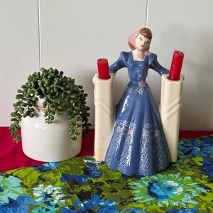 May include: A vintage ceramic candle holder featuring a woman in a blue dress and pink headscarf. The holder has two white columns with red candles.