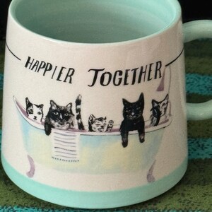 May include: A ceramic mug with a light blue interior and handle, featuring the phrase "Happier Together" in black text. The mug showcases a whimsical illustration of cats in a bathtub, with a light blue and purple color scheme.
