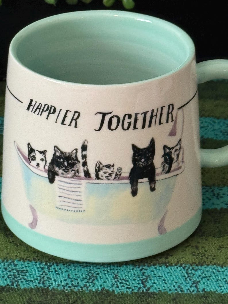 May include: A ceramic mug with a light blue interior and handle, featuring the phrase "Happier Together" in black text. The mug showcases a whimsical illustration of cats in a bathtub, with a light blue and purple color scheme.