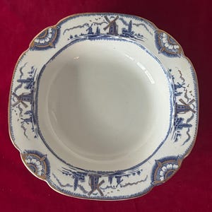 May include: A white, scalloped-edge serving bowl with a blue and gold windmill design around the rim. The bowl has a wide, shallow shape and a gold trim. The interior is plain white.