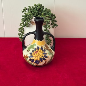 May include: A decorative ceramic vase with a black neck and handles, and a cream-colored body. The vase features a floral design with yellow, blue, and green accents. The vase is displayed on a red surface.