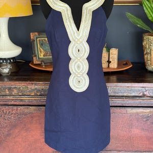 Pu&ograve; includere: Un abito senza maniche blu navy con scollo a V e finiture decorative oro e bianco. Le finiture sono presenti intorno allo scollo e sulla parte anteriore dell'abito. L'abito &egrave; esposto su un manichino nero.
