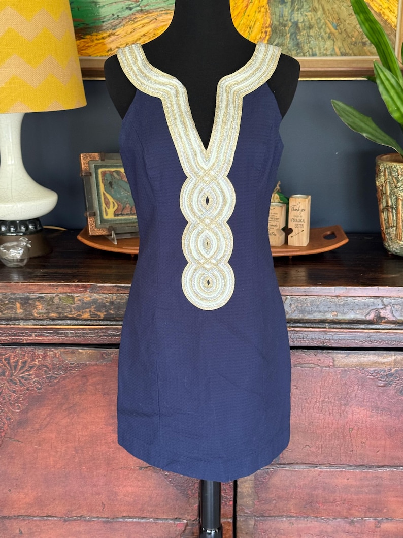 Pu&ograve; includere: Un abito senza maniche blu navy con scollo a V e finiture decorative oro e bianco. Le finiture sono presenti intorno allo scollo e sulla parte anteriore dell'abito. L'abito &egrave; esposto su un manichino nero.