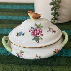 May include: A white porcelain lidded tureen featuring a small sculpted bird finial on the lid. The tureen is decorated with hand-painted floral motifs in pink, blue, and orange with green leaves. It has two light green handles and gold trim.