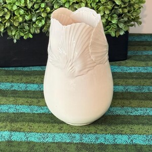 May include: A white ceramic vase with a smooth, rounded base and a top rim sculpted to resemble overlapping flower petals. The surface of the vase features a glossy finish, highlighting the delicate, curved textures of the petal-like design.