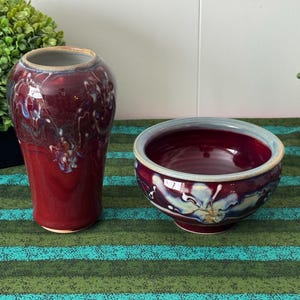 May include: A pair of handmade ceramic pieces, a vase and a bowl, in a rich burgundy color with white and blue accents. The vase is tall and cylindrical, while the bowl is round with a light blue rim. Both pieces sit on a green and blue striped surface.