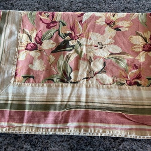 May include: A pink and white floral tablecloth with a green and brown striped border. The tablecloth has a floral pattern with large white flowers and smaller pink flowers.