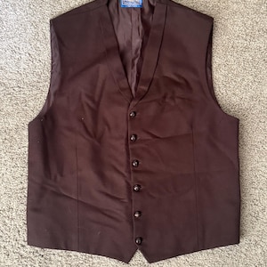 May include: A dark brown vest featuring a V-neckline and a front button closure with six matching buttons. The garment includes a blue Pendleton brand label stitched at the inner neckline. The design is sleeveless with a straight hemline.