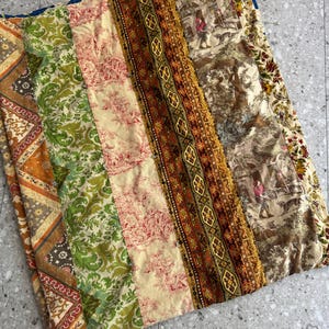 May include: A patchwork quilt featuring vertical strips of fabric in various patterns and colors, including green damask, red and cream toile, brown geometric prints, and a brown and cream scene depicting figures and dogs in a landscape.
