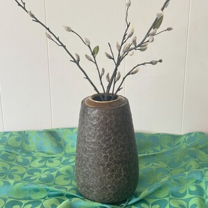 May include: A dark brown textured vase with a bronze rim holds branches with small, white, and green buds. The vase sits on a green and blue patterned fabric. The background is a white wall.