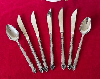 May include: A set of seven silver-plated utensils, including five butter knives and two spoons, with ornate handles. The utensils are arranged on a red surface.