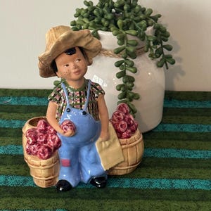 May include: A ceramic figurine of a young boy in blue overalls and a straw hat, holding a red fruit and a cloth. Two small wooden barrels filled with red fruit flank him. A white vase with green plants is in the background.