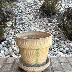 May include: A weathered, beige ceramic flower pot with a matching saucer. The pot features a repeating arched design and subtle green and blue accents. The pot sits on a brick surface, with a background of pebbles and greenery.