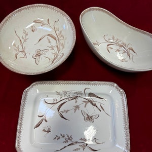 May include: Three white ceramic dishes with a brown floral pattern featuring butterflies and grasses. The dishes include a round bowl, an oval bowl, and a rectangular serving platter.