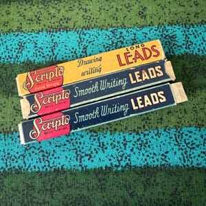 May include: Three rectangular Scripto lead refill boxes stacked vertically. The top box is yellow with red and black text reading Long Leads and Drawing writing. The two bottom boxes are dark blue with white text reading Smooth Writing Leads.