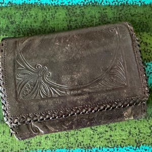 May include: A dark brown leather wallet featuring an embossed decorative pattern on the front flap. The edges of the wallet are finished with a braided leather lacing detail that runs along the perimeter of the rectangular folding structure.