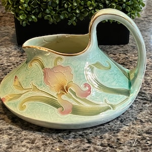 May include: A light blue ceramic pitcher with a gold rim and a floral design featuring a pink and yellow iris. The pitcher has a wide base and a curved handle.