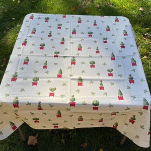 Può includere: Una tovaglia color crema con un motivo ripetuto di cactus verdi in vaso e piccole foglie verdi. I cactus sono in vasi rossi. La tovaglia è drappeggiata su un tavolo quadrato, con i bordi che pendono.