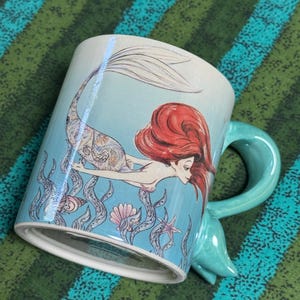 May include: A light blue ceramic mug featuring a detailed illustration of a mermaid with long red hair swimming among seaweed, seashells, and a starfish. The mug has a unique handle shaped like a mermaid tail, extending from the side of the vessel.