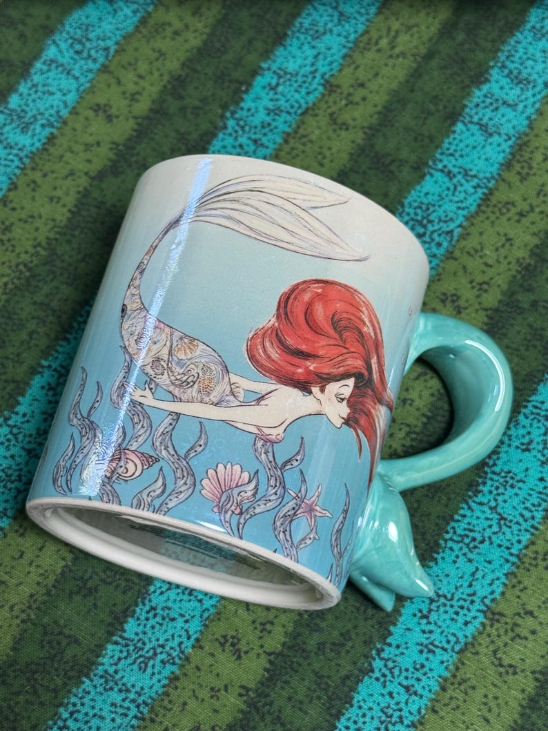 May include: A light blue ceramic mug featuring a detailed illustration of a mermaid with long red hair swimming among seaweed, seashells, and a starfish. The mug has a unique handle shaped like a mermaid tail, extending from the side of the vessel.