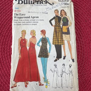 May include: Vintage Butterick sewing pattern for a wraparound apron, dress, or tunic. The pattern features illustrations of the finished garments, including a long red dress and a plaid apron. Sizes include petite, small, and medium.